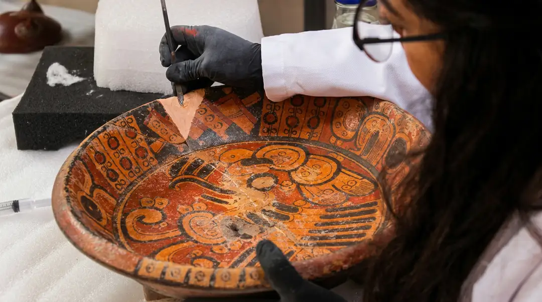 Tulum’s Museum Just Received a Jar Older Than the Walled City Itself A conservator wearing black gloves and glasses carefully cleans an ancient ceramic bowl decorated with orange and black geometric patterns and animal motifs