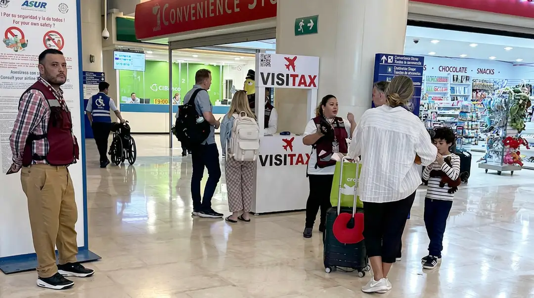 Tourists and staff interact at a visitor information booth in an airport terminal with duty-free shops visible in the background