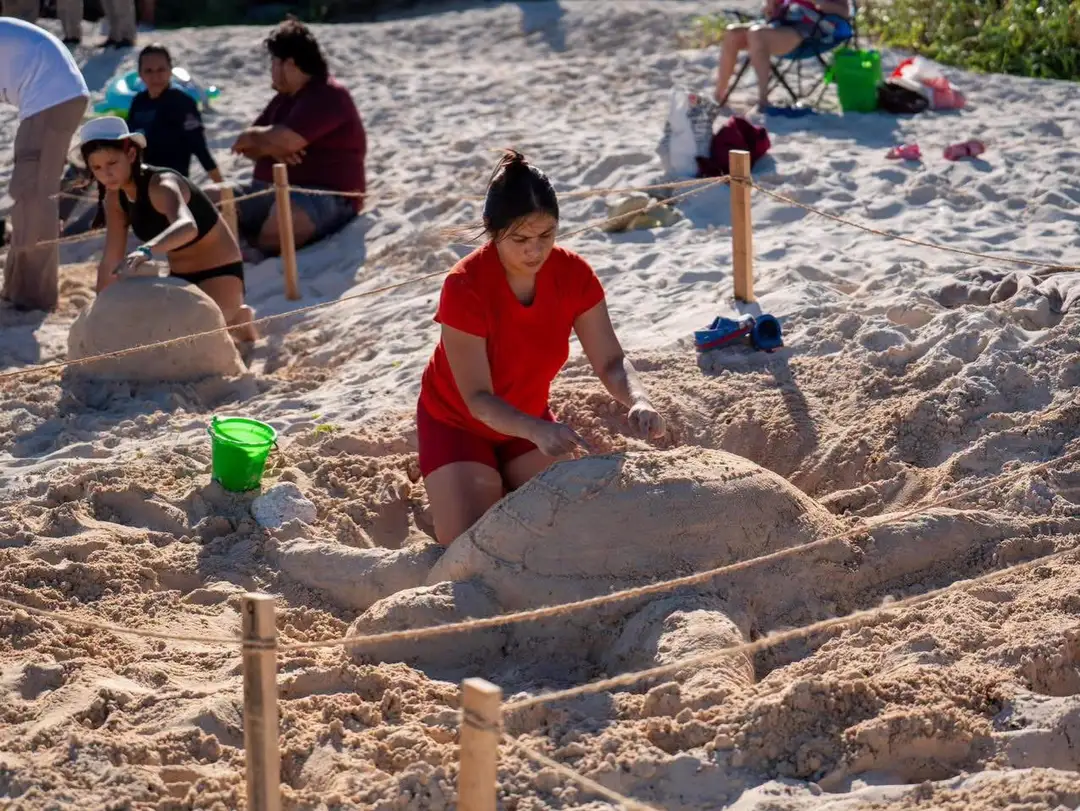 Girl in red shirt sculpts sand on a beach while other participants work on sand sculptures nearby