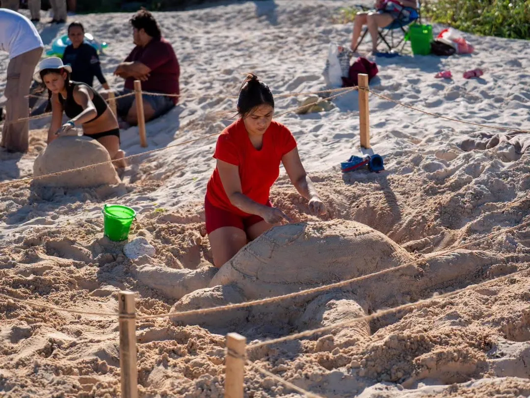 Girl in red shirt sculpts sand on a beach while other participants work on sand sculptures nearby