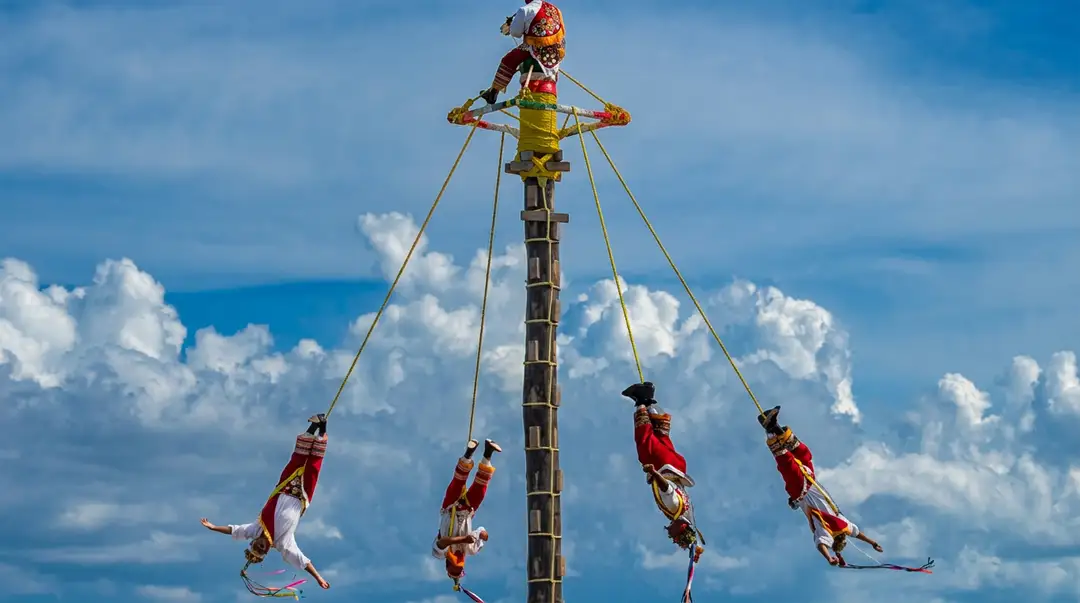 Five performers in traditional red and white clothing swing from ropes attached to a tall wooden pole against a blue sky with white clouds