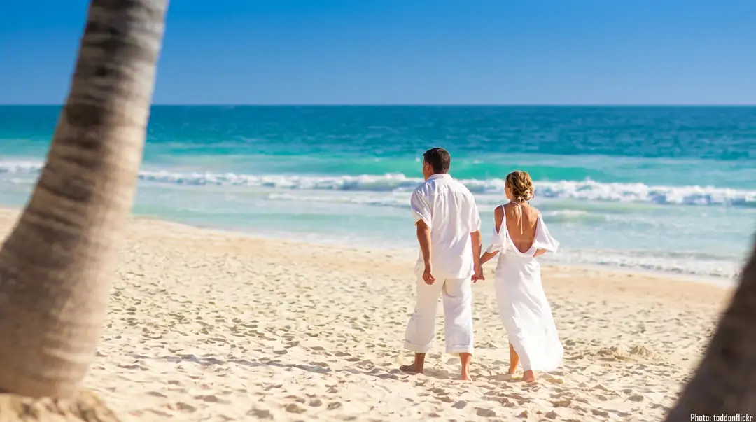 Couple in white clothing holding hands on a sandy beach with ocean and waves visible behind them