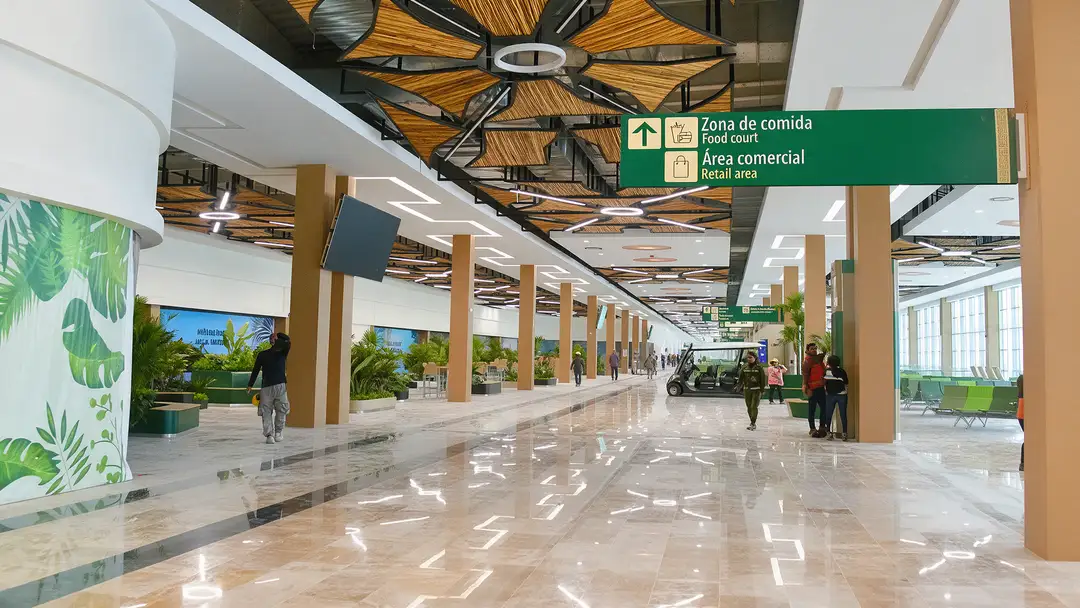 A spacious, modern airport terminal interior with polished floors, wooden ceiling elements, support columns, directional signage, and scattered pedestrians walk