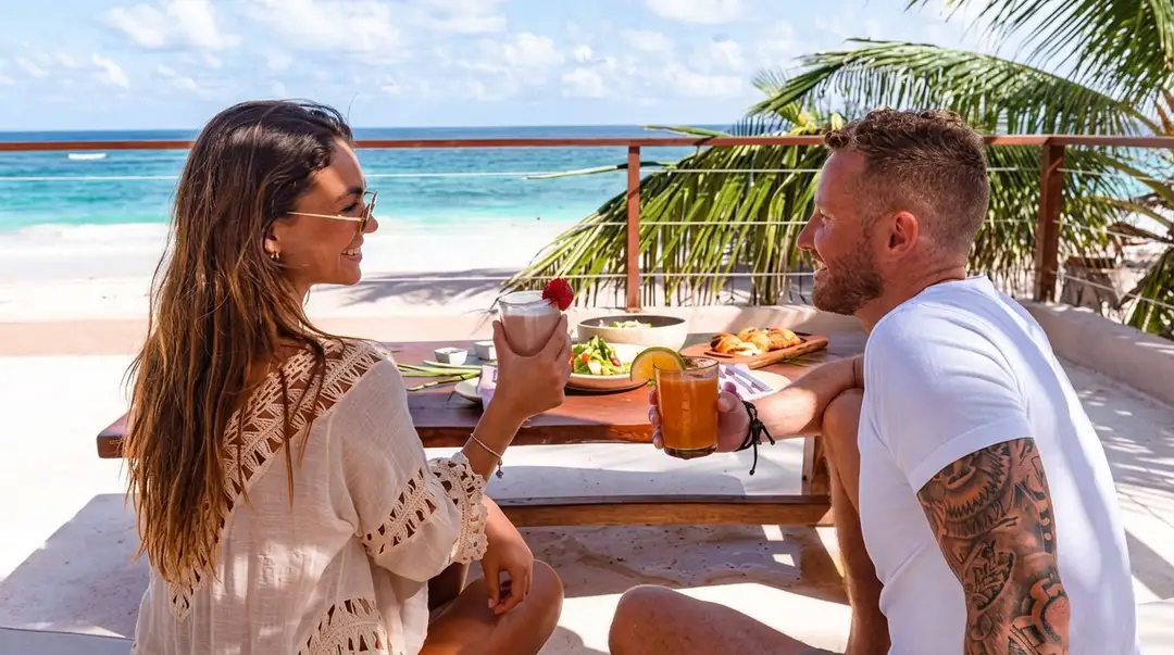 Why Overcharging This Winter Could Be the Most Expensive Mistake Tulum Businesses Make Two people sit at a beachfront table with food and drinks, ocean and palm trees visible behind them