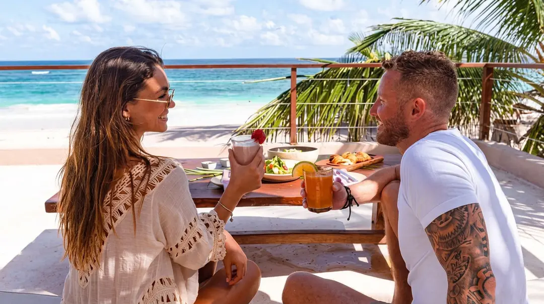 Why Overcharging This Winter Could Be the Most Expensive Mistake Tulum Businesses Make Two people sit at a beachfront table with food and drinks, ocean and palm trees visible behind them