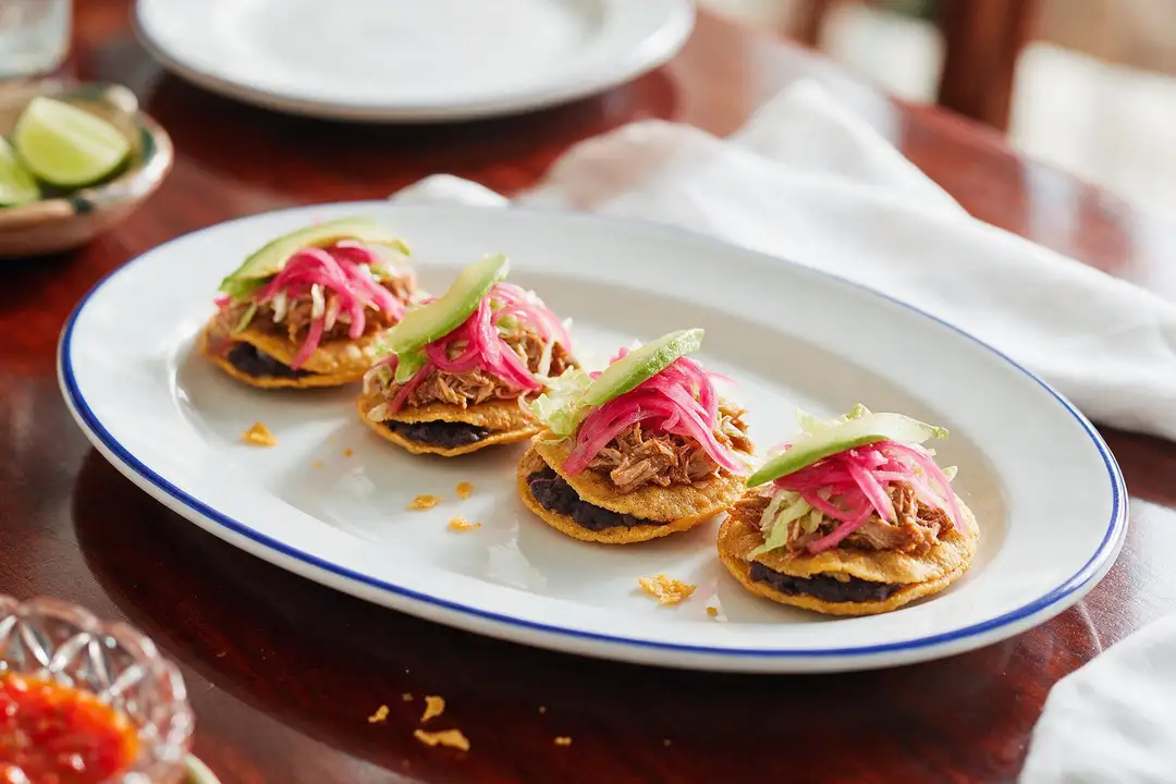 Panuchos: Yucatecan bean-stuffed tortillas, fried and topped Four fried tortillas stuffed with black bean paste, topped with shredded pork, pickled red onion and avocado, on a cobalt rimmed platter